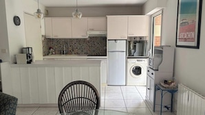 A compact kitchen with white cabinets, a built-in washing machine, and a dining area with a chair and table. - Louisiana apartment (Arcachon)