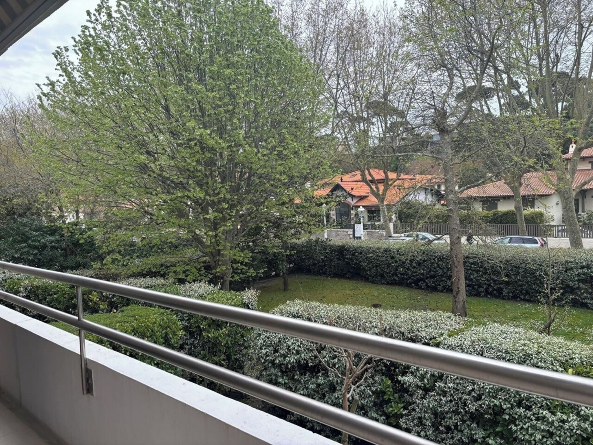 A balcony with a metal railing overlooking a residential area with houses, trees, and a well-maintained lawn.