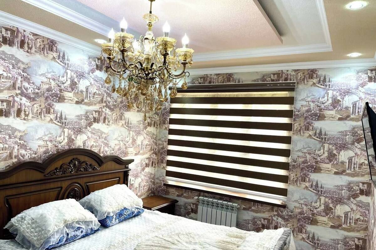 A bedroom with a large bed, a chandelier, and a window with blinds.