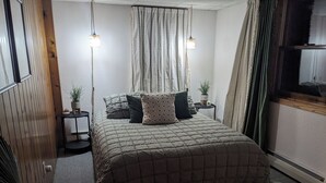Un dormitorio con una cama, dos mesitas de noche, una lámpara colgante, una planta en maceta y una ventana con cortinas.