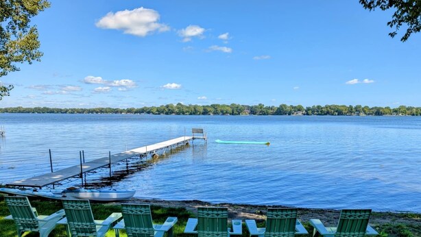 Vista al lago con un muelle que se adentra en el agua, rodeado de sillas verdes y una mesa de picnic.