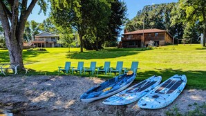 Kayaks alineados en un césped cerca de un cuerpo de agua, con un edificio y árboles al fondo.