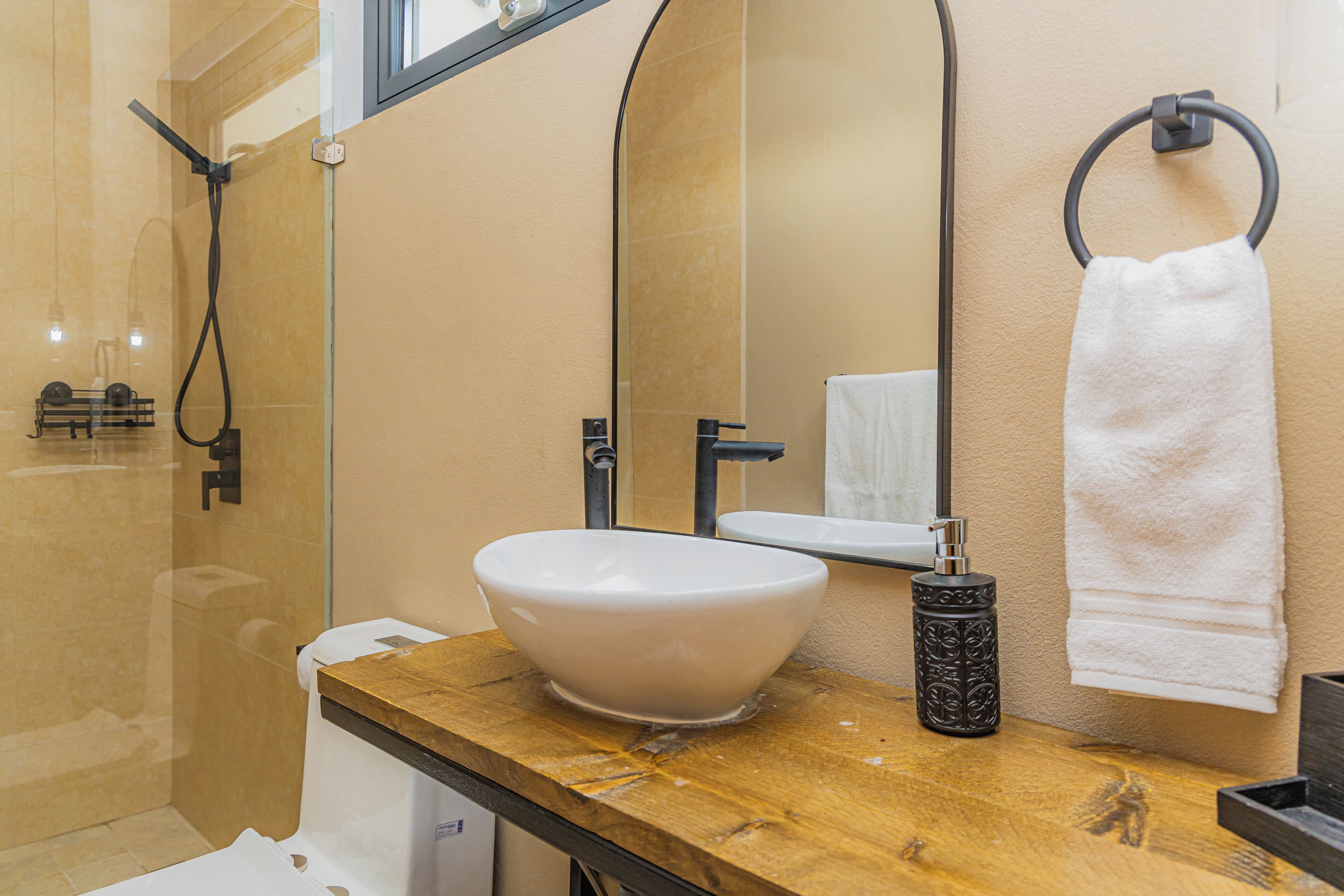 A bathroom with a large mirror, a white sink, a wooden countertop, a towel ring, and a shower area.
