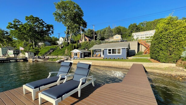 A wooden deck with lounge chairs by a calm water body, surrounded by greenery and houses. - Swim, Sip, and Stay Awhile at this Beautiful Keuka Lake Retreat! (Dundee)
