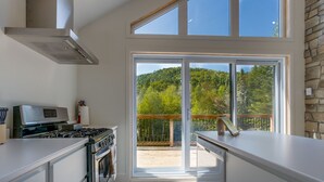 A modern kitchen with a gas stove, oven, and a large window overlooking a deck with a forest view.