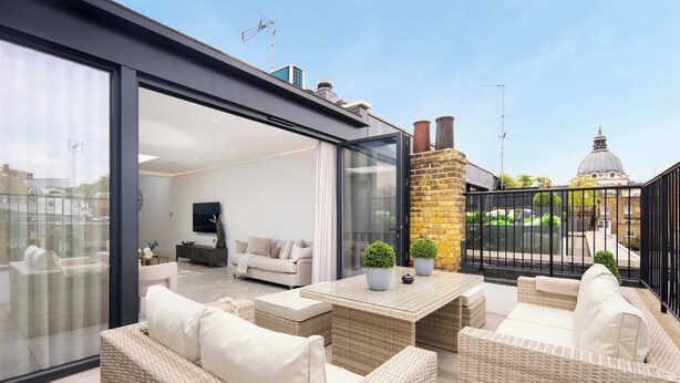 A modern outdoor living space with wicker furniture, a glass door leading to an interior room, and a view of a dome structure in the background.