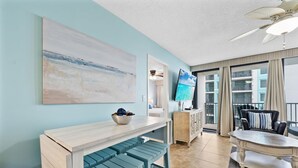A modern living room with a light blue wall, a large abstract painting, a dining area with a white table and blue benches, and a ceiling fan. - Island Winds West 471 (Gulf Shores)