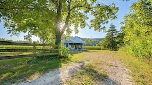 A rural property with a gravel driveway leading to a house with a porch, surrounded by lush greenery and trees. - Hot Tub & Creek Views: Secluded Perryville Cabin (Perryville)