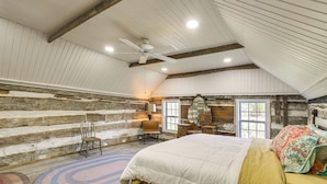 A bedroom with a sloped ceiling, a bed with a white and yellow bedspread, a wooden chair, a round rug, and a wooden wall with a window. - Hot Tub & Creek Views: Secluded Perryville Cabin (Perryville)