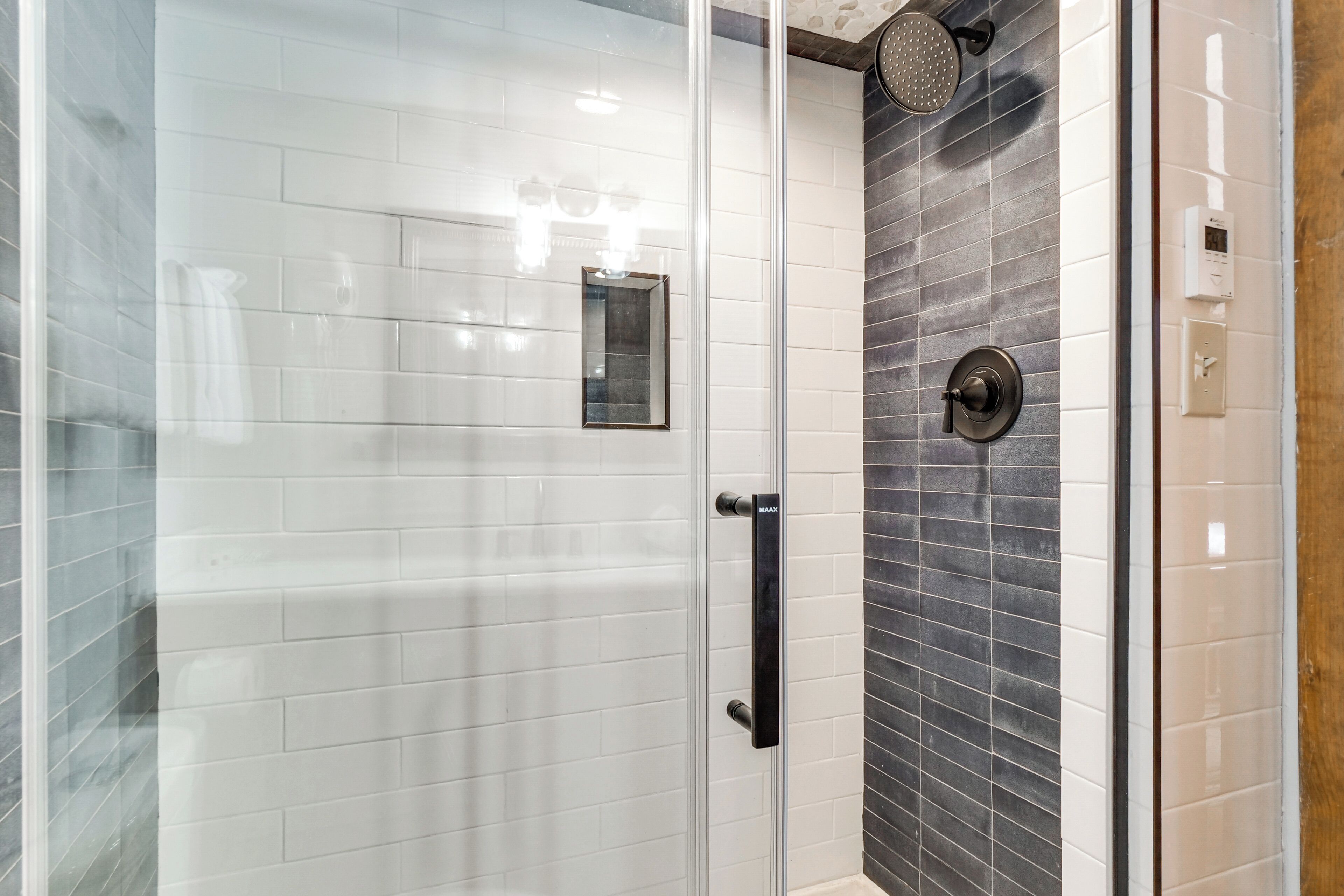 Une douche fermée par des parois de verre, avec des carreaux blancs et gris foncé, une poignée noire et une pomme de douche.