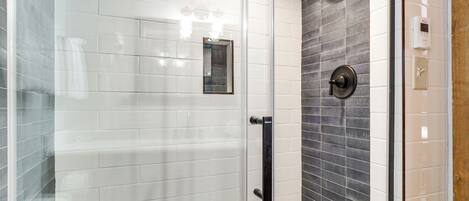 Une douche fermée par des parois de verre, avec des carreaux blancs et gris foncé, une poignée noire et une pomme de douche.