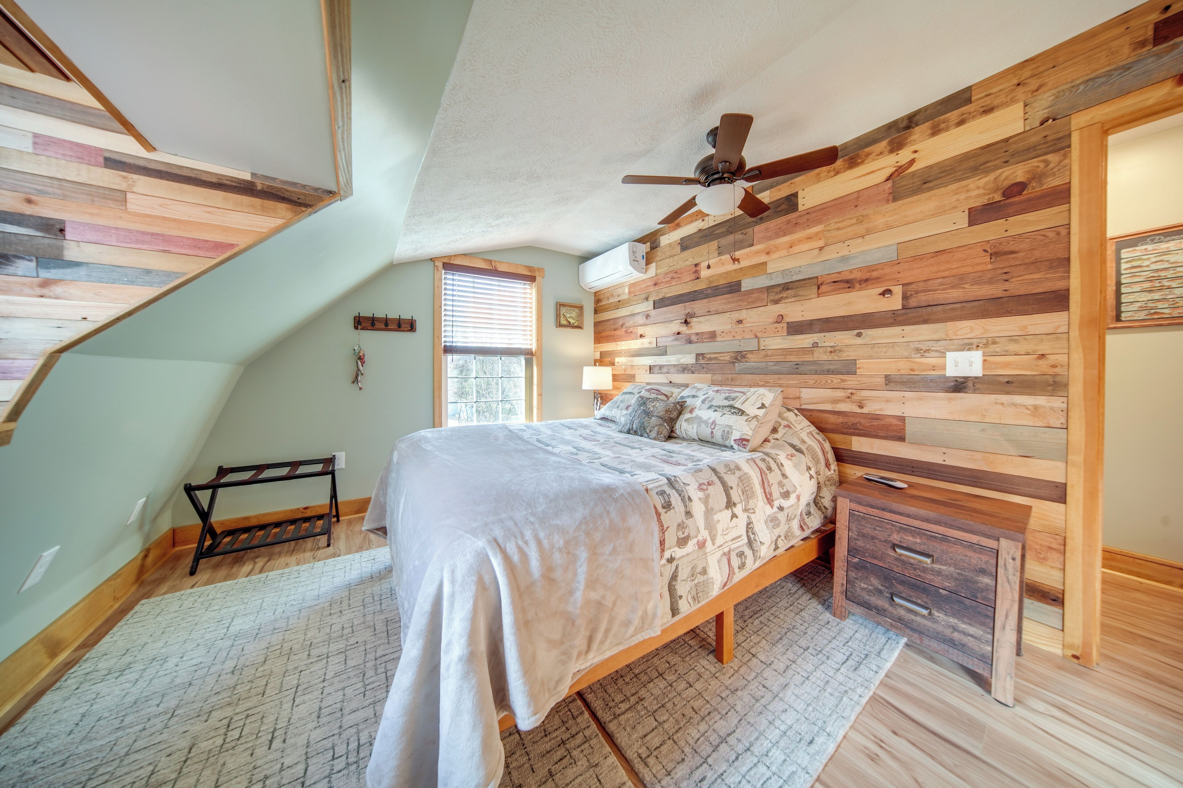 A bedroom with a sloped ceiling, wooden panel wall, a bed with a patterned bedspread, a nightstand, and a ceiling fan.