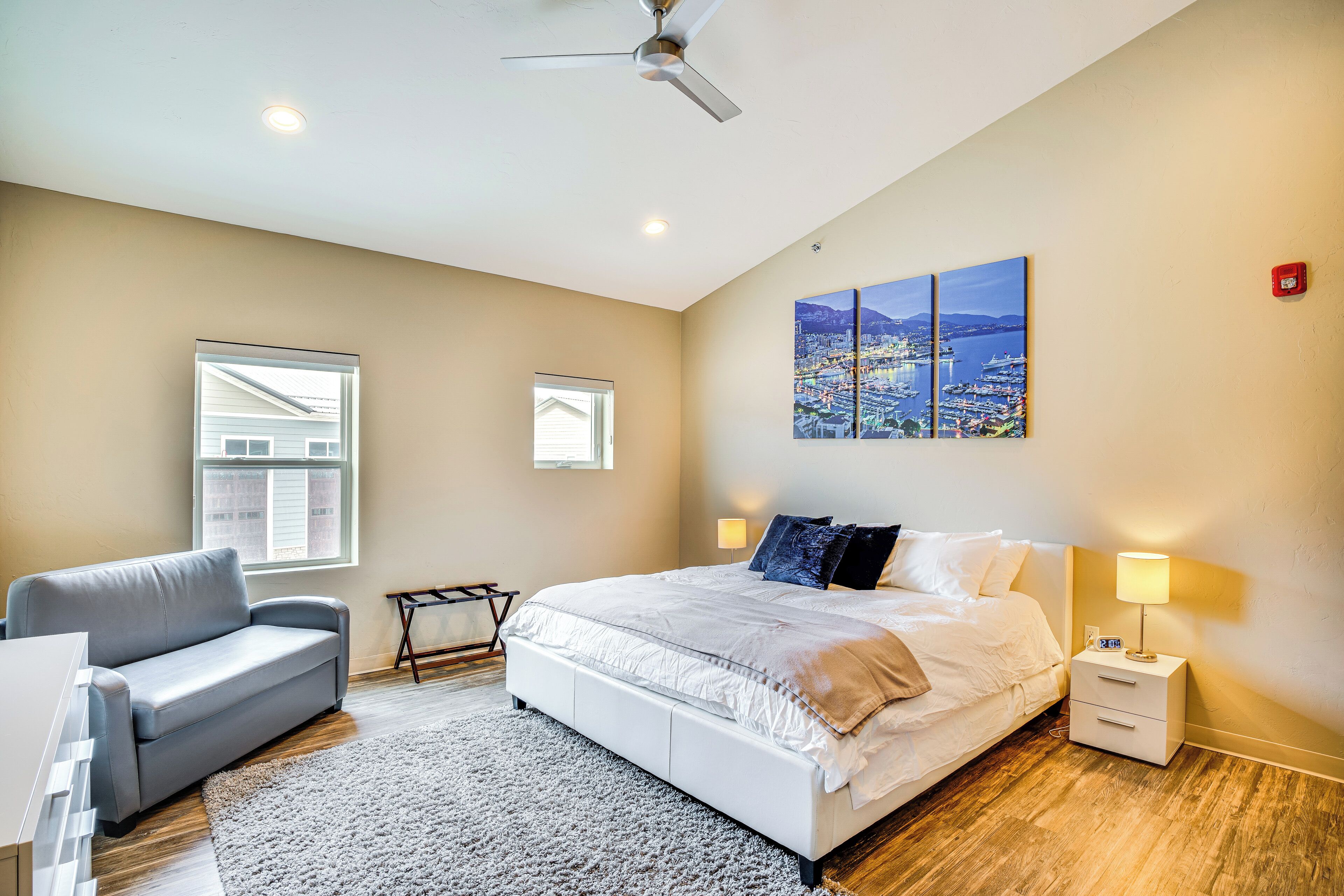 A bedroom with a bed, a gray sofa, a small table, and a nightstand with a lamp.