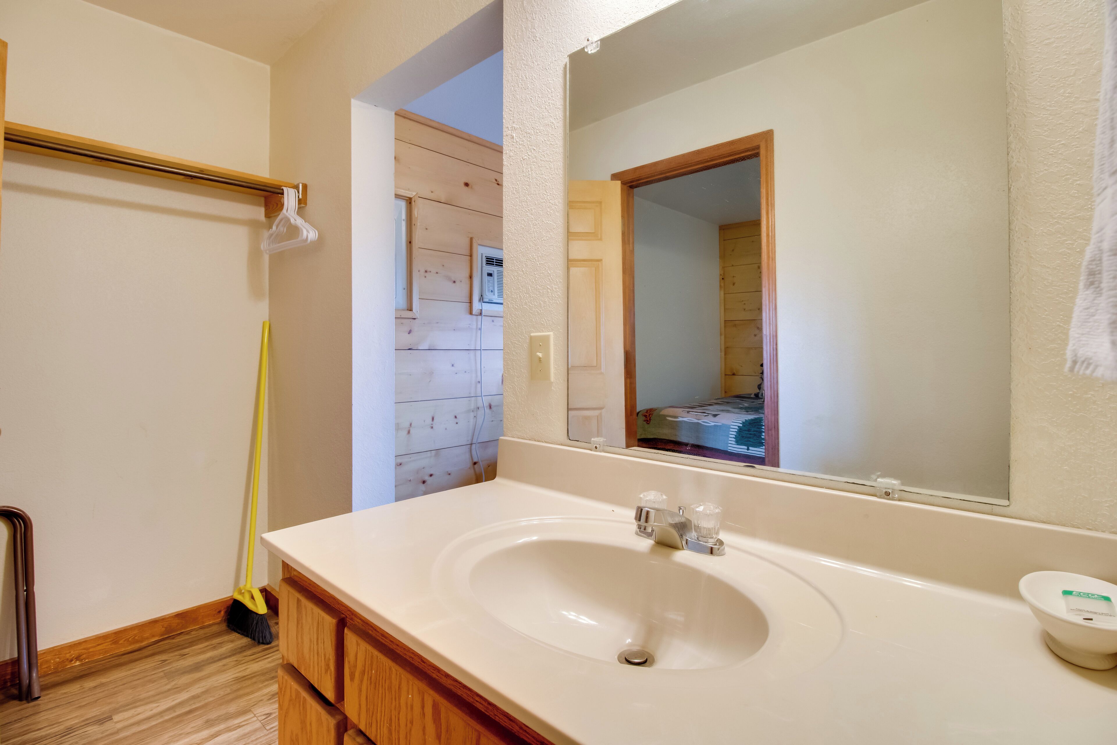 Une salle de bain avec un lavabo blanc, des meubles en bois, un miroir et un balai appuyé contre le mur.