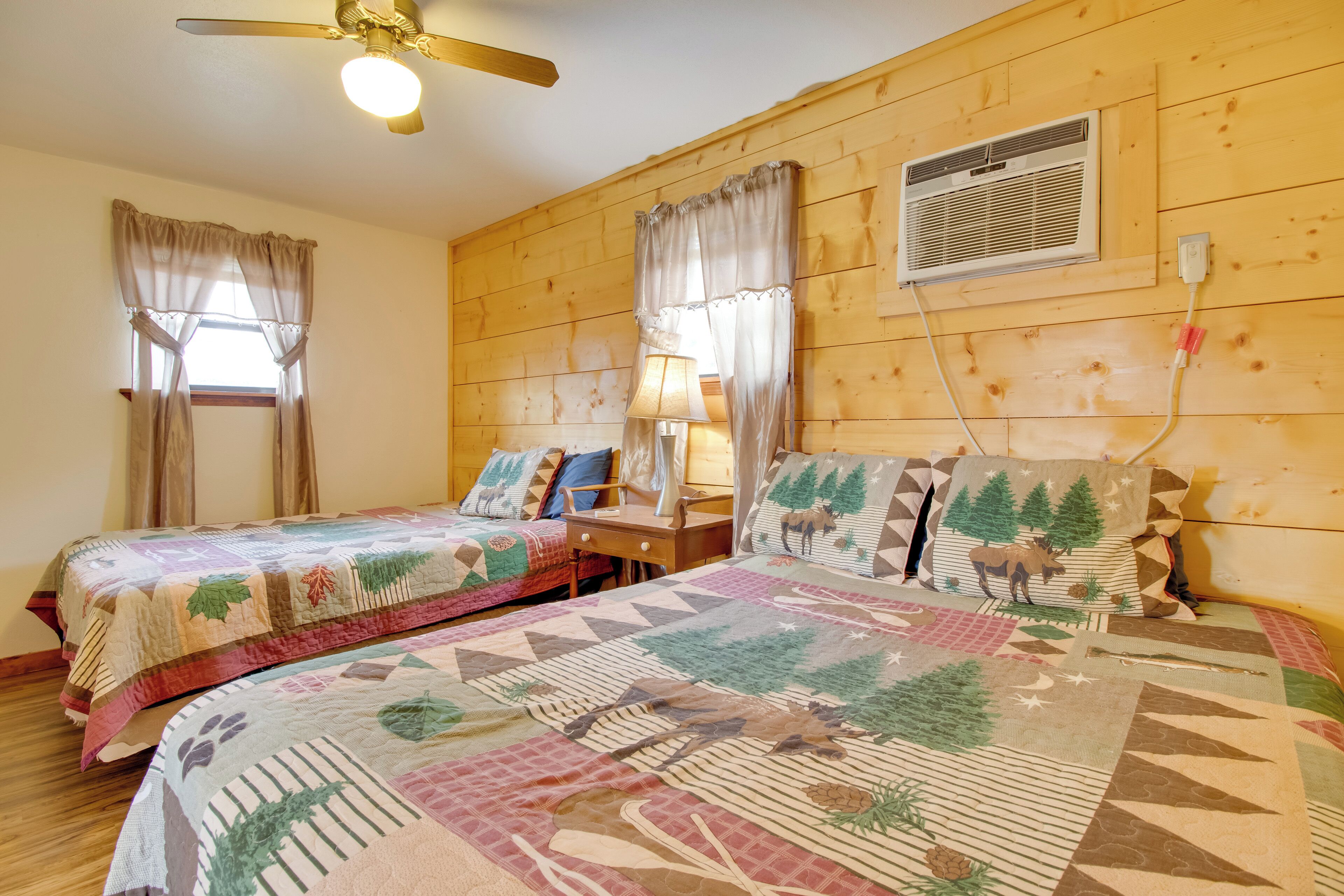 A room with two beds, wooden walls, a ceiling fan, a window with curtains, a desk, and a lamp.