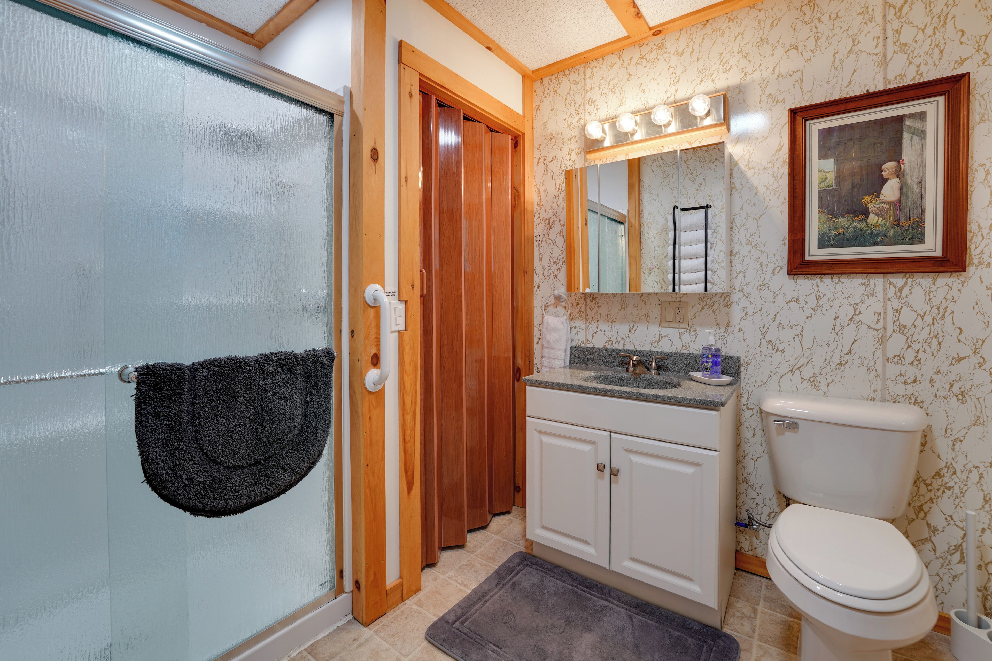 A bathroom with a glass door, wooden door frame, white cabinets, a white toilet, a sink with a mirror above it, and a framed picture on the wall.