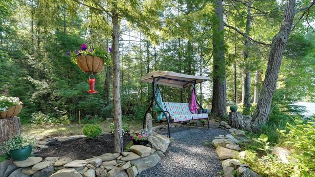 A forested area with a swing set, a hanging flower basket, and a small garden bed with rocks and plants. - Private Dock & Deck: Tranquil Dam Pond Cottage! (Augusta)