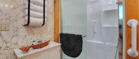 Un baño con ducha de vidrio, un lavamanos blanco y un cesto de artículos de tocador, y un perchero con una toalla blanca.