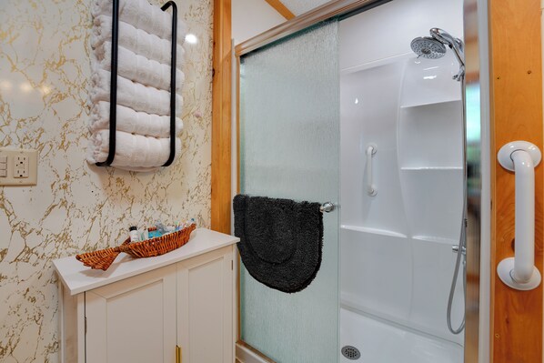A bathroom with a glass-enclosed shower, a white sink with a basket of toiletries, and a towel rack with a white towel.
