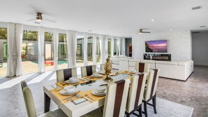 A modern dining area with a table set for four, a sofa, and a TV mounted on a white brick wall. - Huge Family Retreat w/ Heated Pool & Games (Westlake)