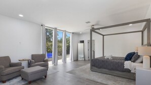 A modern bedroom with a large bed, a sitting area with two armchairs, and a view of the outdoors through large windows. - Huge Family Retreat w/ Heated Pool & Games (Westlake)