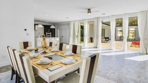 A modern dining area with a glass table, white chairs, and a kitchen in the background. - Huge Family Retreat w/ Heated Pool & Games (Westlake)