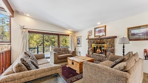A cozy living room with a fireplace, comfortable seating, and large windows offering a view of the outdoors. - Spacious 4,000 Sq Ft Riverfront Home | Game Room • Deck • 1.5 Miles to Keystone (Keystone)