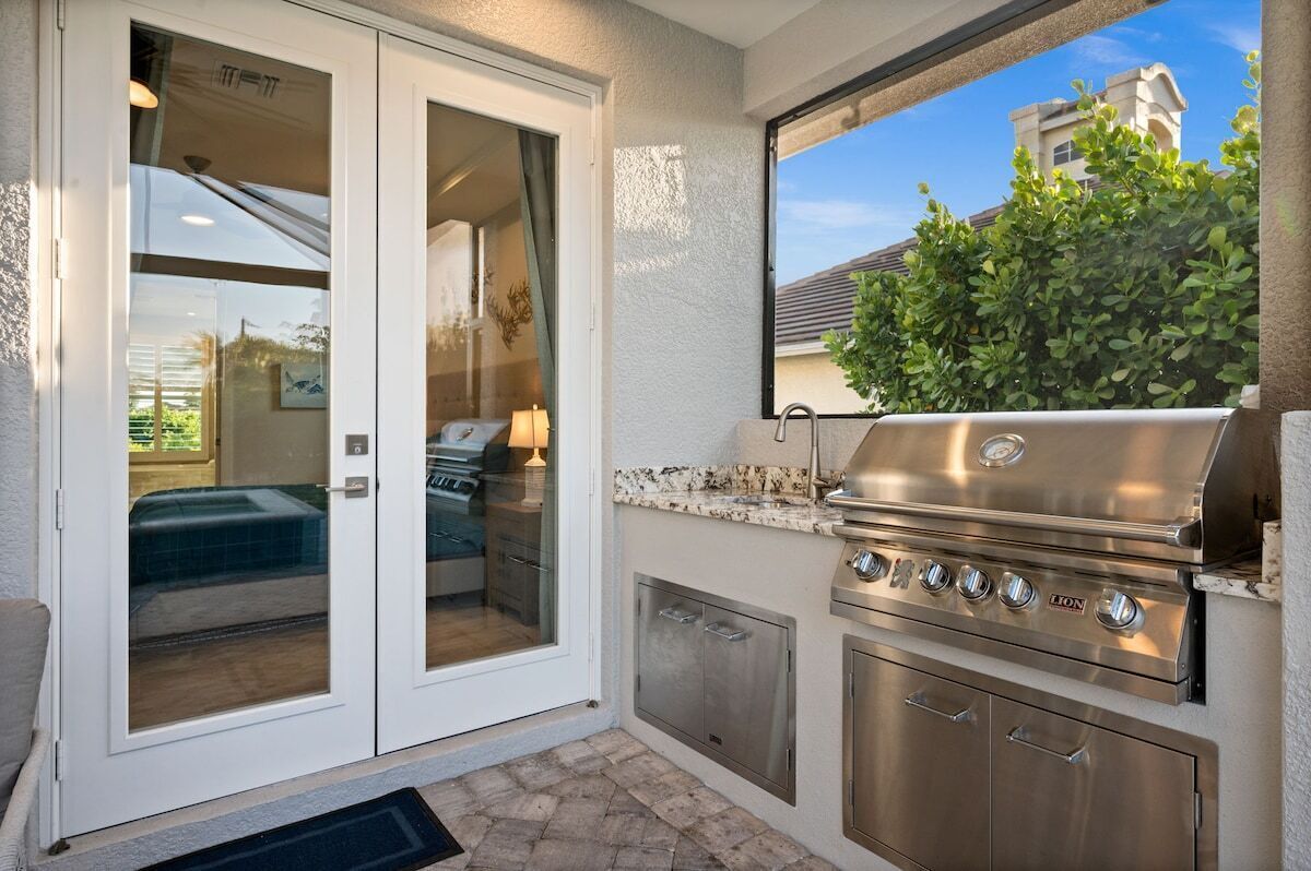 An outdoor kitchen with a stainless steel grill, a sliding glass door leading to a patio, and a view of a building in the background.
