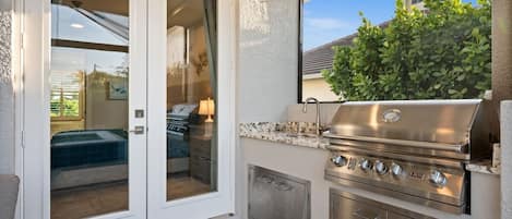 An outdoor kitchen with a stainless steel grill, a sliding glass door leading to a patio, and a view of a building in the background.