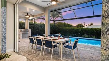An outdoor dining area with a pool view, a ceiling fan, and a kitchen in the background.