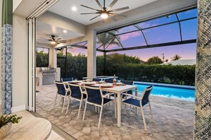 An outdoor dining area with a pool view, a ceiling fan, and a kitchen in the background. - Modern Luxury • 7 Min to Beach • Heated Pool & Spa (Marco Island)