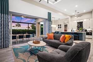 Una sala de estar moderna con sofá, sillones y una mesa de centro. Hay una zona de comedor con sillas y una mesa, y una cocina con armarios y electrodomésticos.