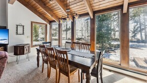 A dining area with a wooden table and chairs, large windows with a snowy landscape view, and a television on the wall. - Spacious Forest-Backed Lodge • 5BR • Game Room • Minutes to Slopes (Keystone)