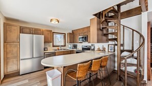 A modern kitchen with wooden cabinets, a stainless steel refrigerator, a large island with bar stools, and a spiral staircase. - Spacious Forest-Backed Lodge • 5BR • Game Room • Minutes to Slopes (Keystone)