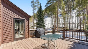 A wooden deck with a glass door leading to a room, a covered grill, and a table with a cushion. - Spacious Forest-Backed Lodge • 5BR • Game Room • Minutes to Slopes (Keystone)