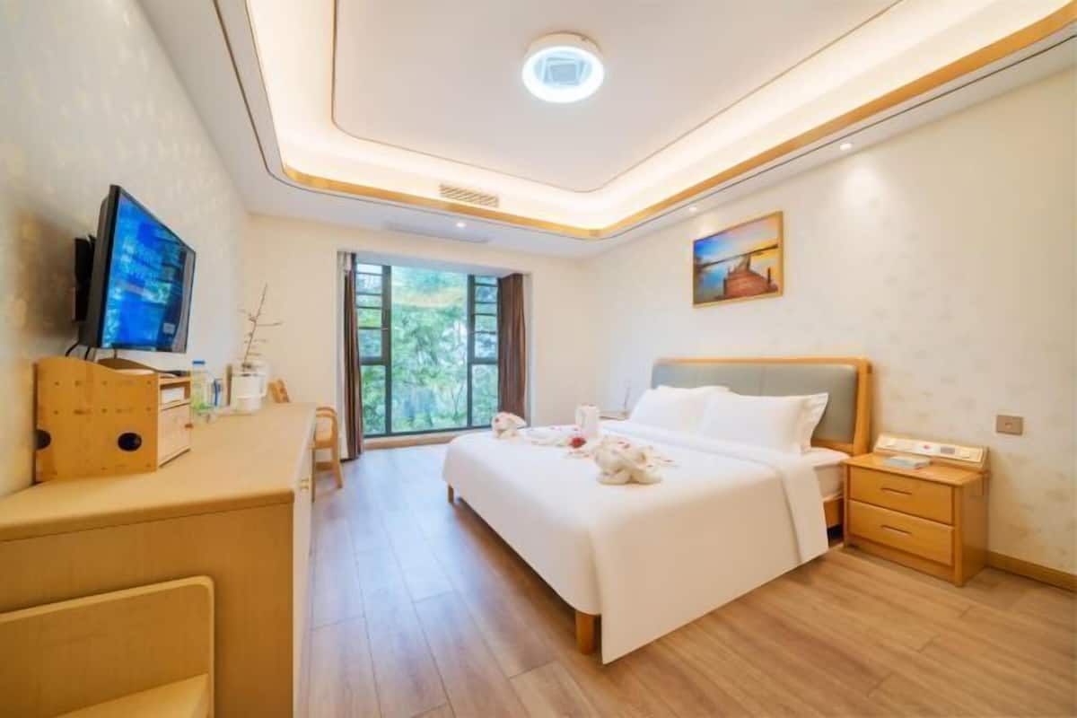 A modern hotel room with a large bed, bedside tables, a desk with a computer, and a window with a view of greenery.