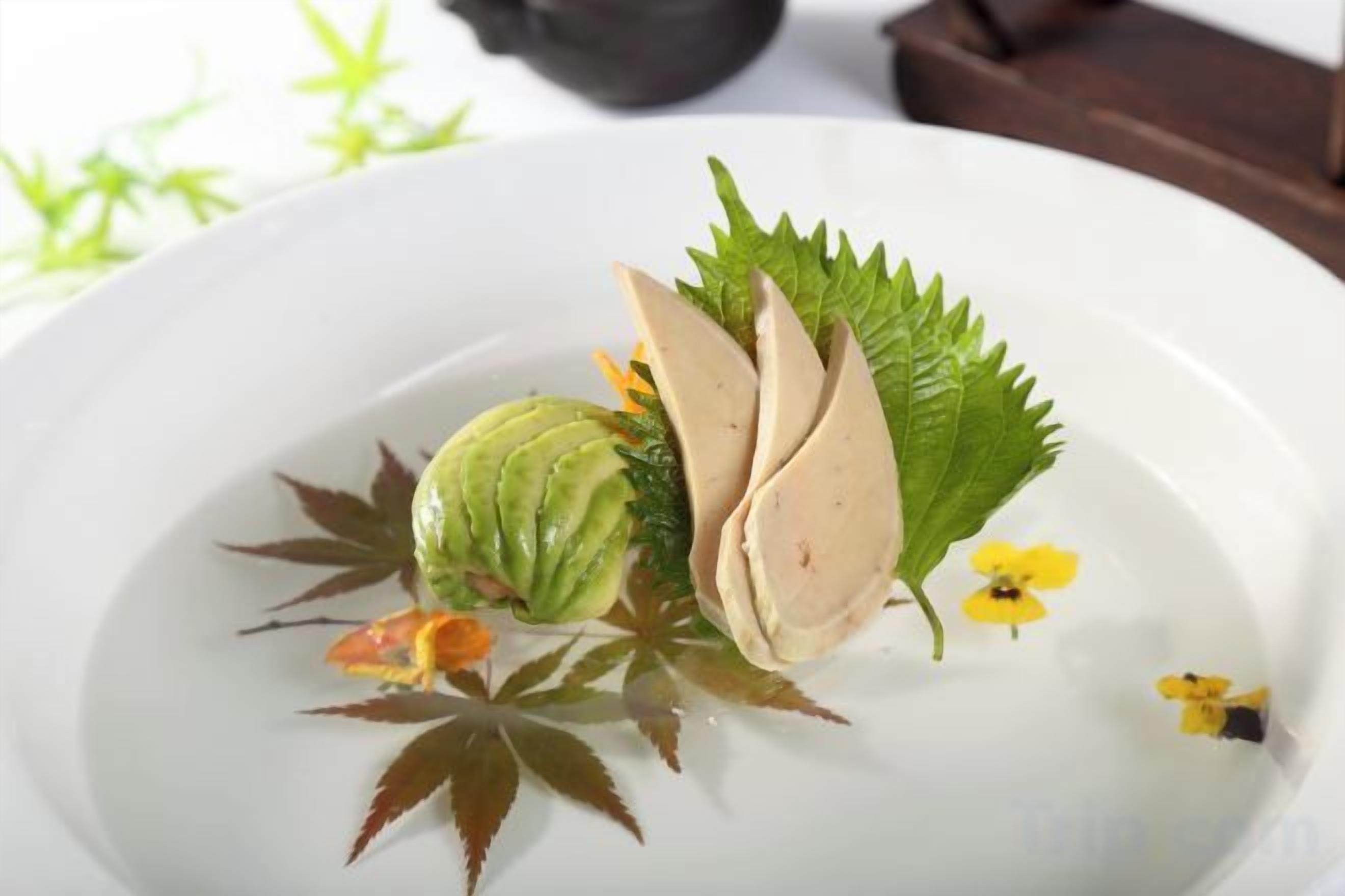 A white plate with a decorative arrangement including a green fruit, leaves, and small flowers.