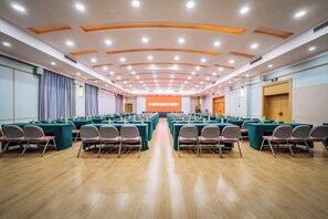Una sala de conferències amb files de taules i cadires, un escenari amb pantalla de presentació i un terra de fusta.