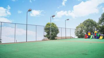 Una cancha de tenis al aire libre con superficie verde, rodeada por una valla metálica, y un parque infantil con estructuras de juego de colores al fondo.