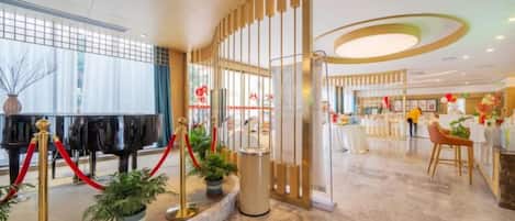 A hotel lobby with a grand piano, red ropes, potted plants, and a reception desk.