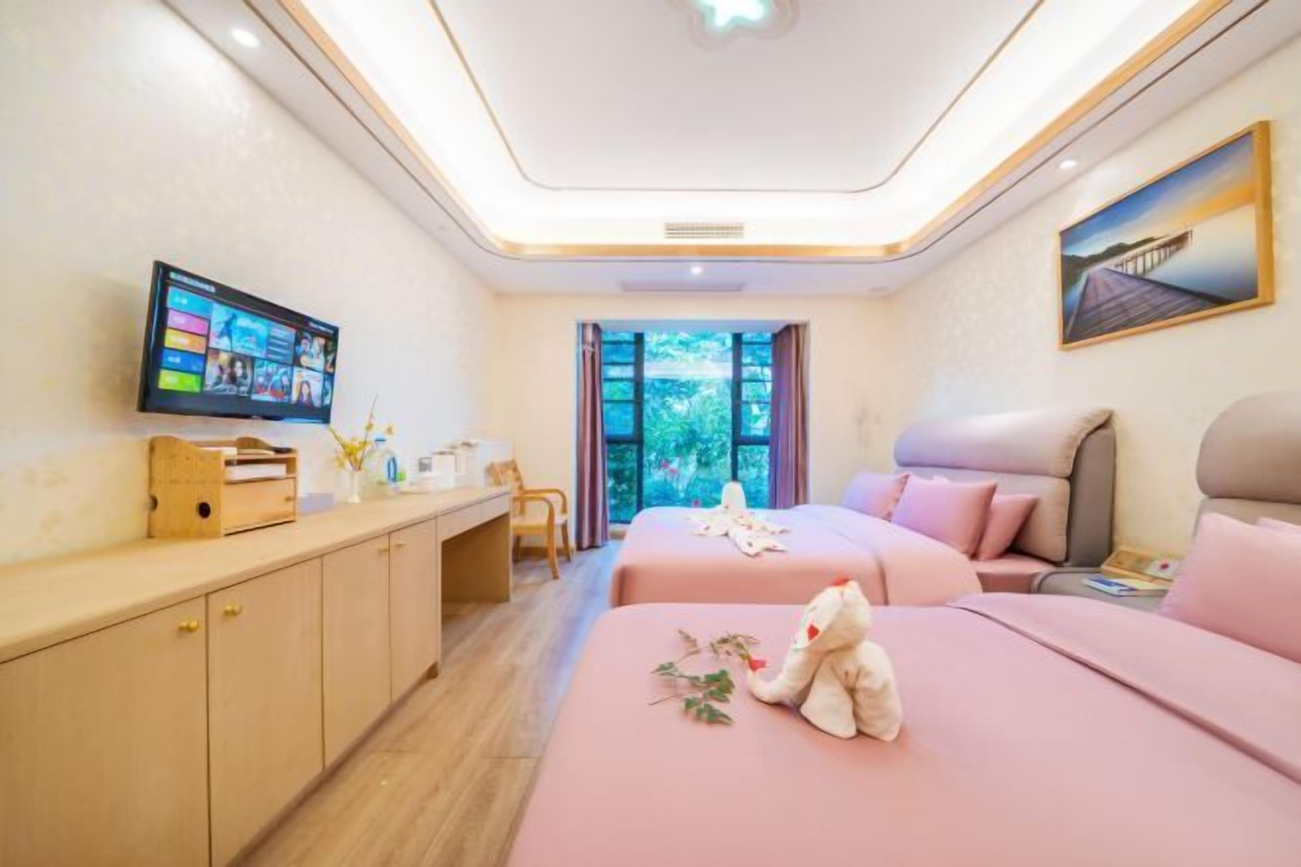 A hotel room with two beds, a desk, a television, and a window with a view of greenery.