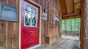 Une cabane en bois avec une porte rouge, une pancarte indiquant «Bienvenue» et un banc à l’extérieur.