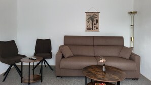 A living room with a brown sofa, two black chairs, a small round table, a floor lamp, and a wall hanging. - Vacation Home with a View of Stolpen Castle at the "Gateway to Saxon Switzerland" (Dürrröhrsdorf-Dittersbach)