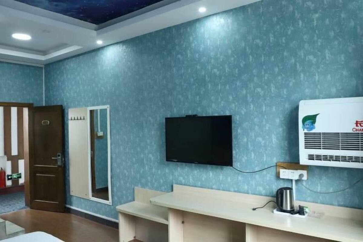 Une chambre d’hôtel avec un mur bleu à texture, un téléviseur à écran plat fixé au-dessus d’un bureau en bois et un climatiseur mural.
