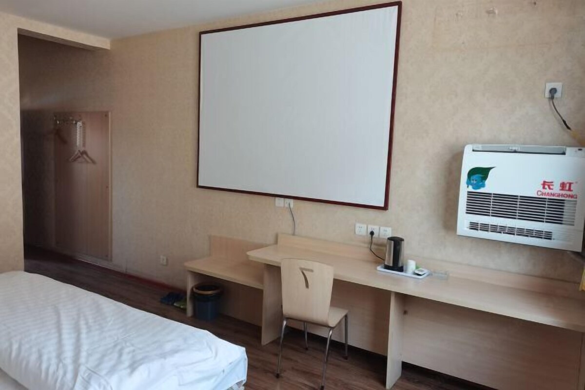 A hotel room with a bed, a desk, a chair, a whiteboard, and an air conditioning unit.