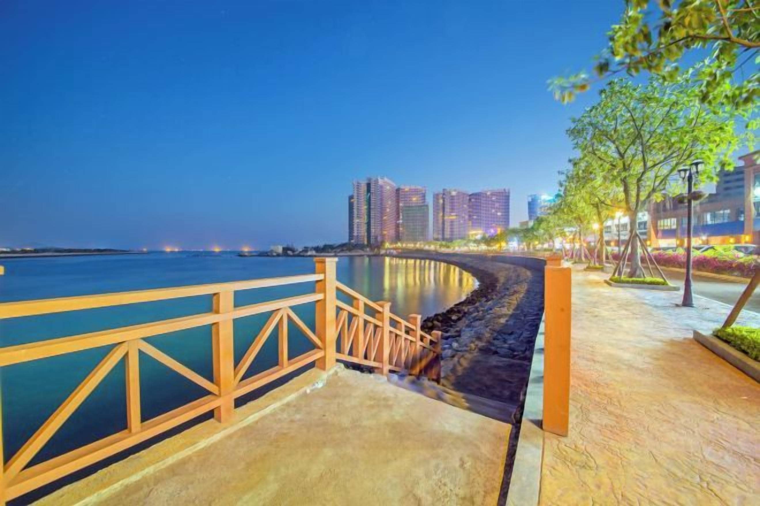A waterfront promenade with a wooden railing, a concrete walkway, and buildings in the background.