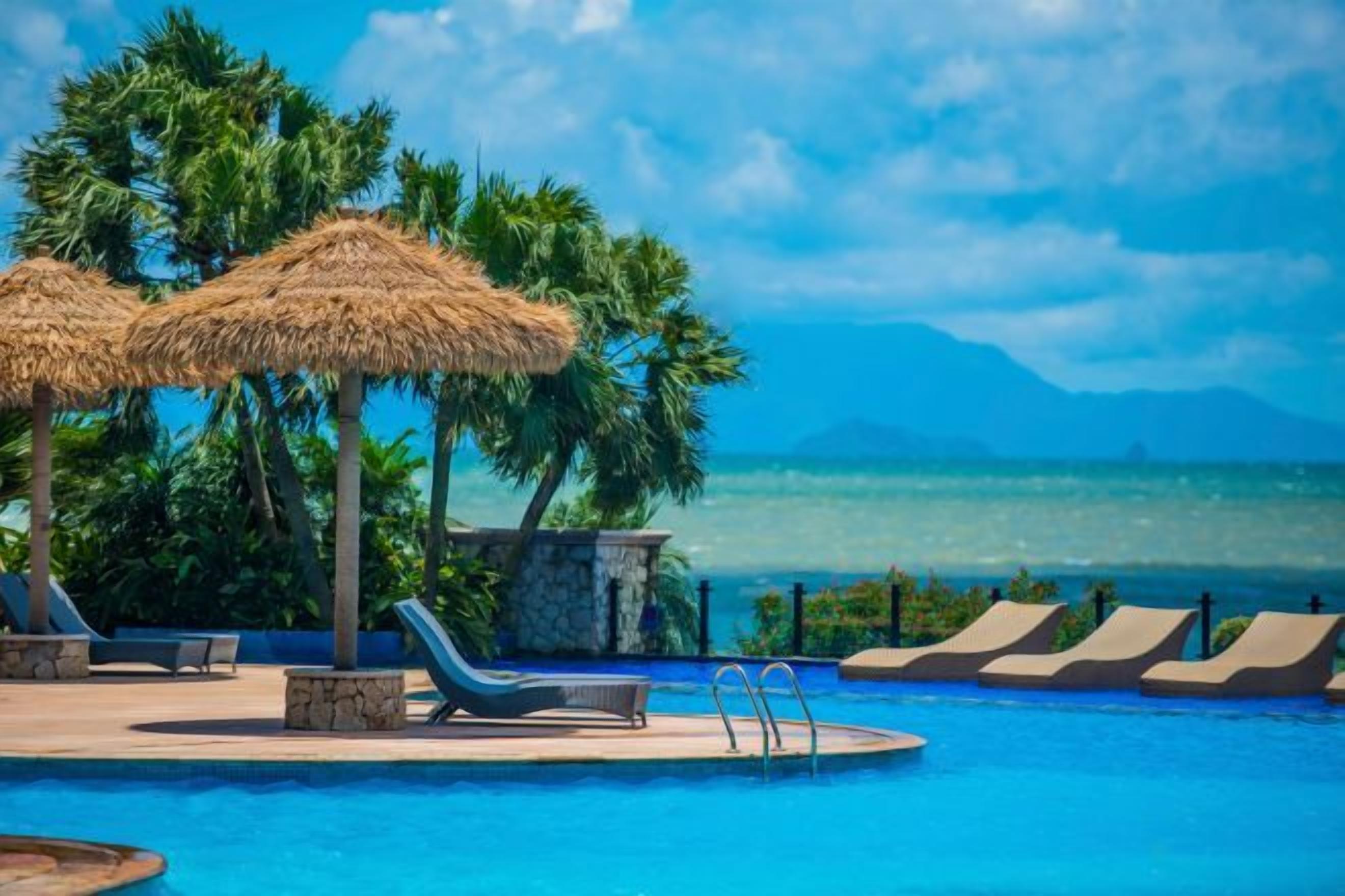 A pool area with lounge chairs and thatched umbrellas, overlooking a body of water and mountains.