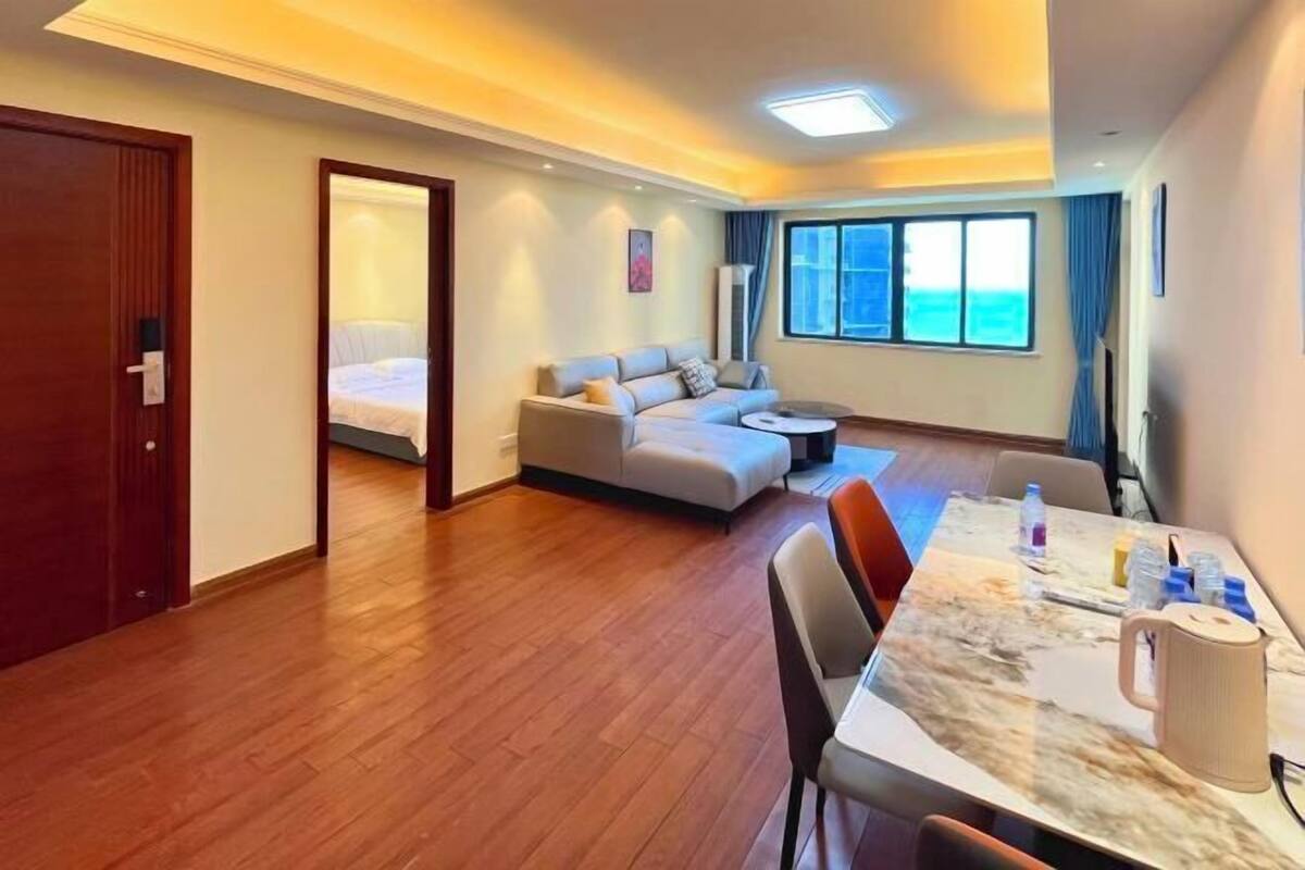 Een hotelkamer met een houten vloer, een bank, een eettafel met stoelen, een bed met uitzicht op zee en een groot raam met gordijnen.