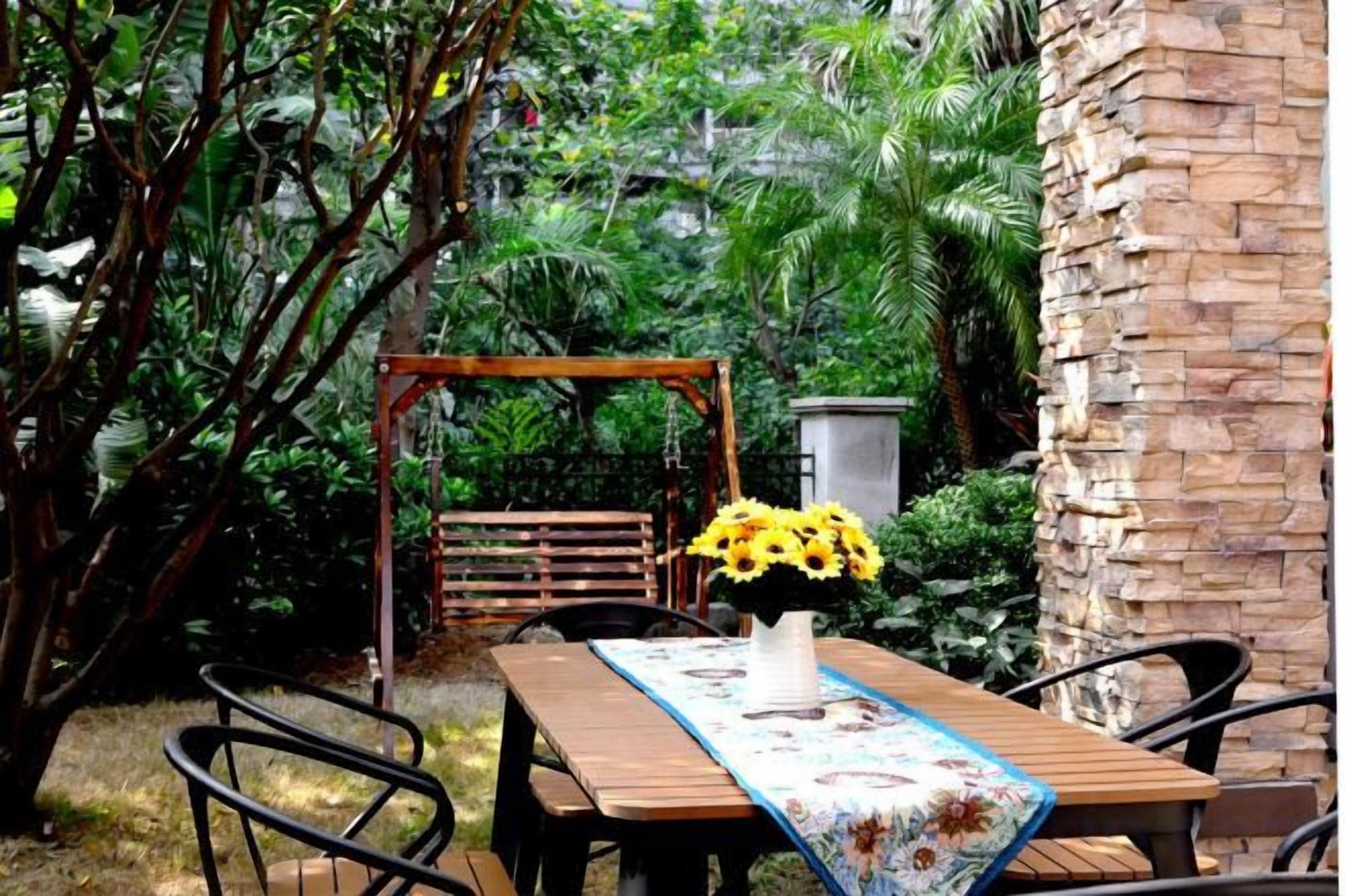 Uma mesa de madeira com toalha de mesa florida, rodeada por cadeiras, num jardim com baloiço e um muro de pedra.
