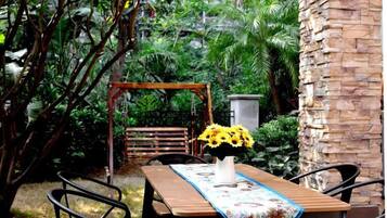 Uma mesa de madeira com toalha de mesa florida, rodeada por cadeiras, num jardim com baloiço e um muro de pedra.